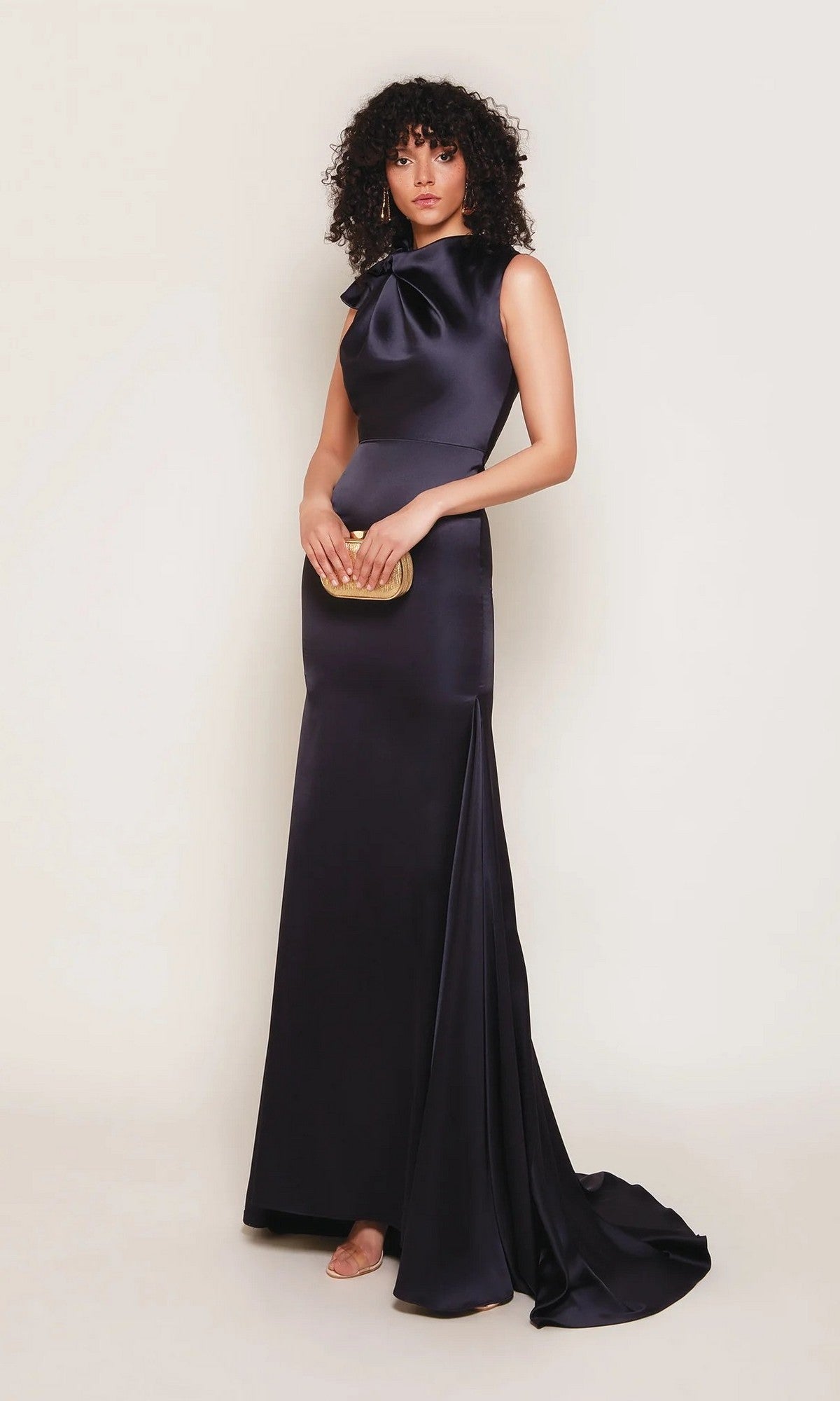 A woman in the Alyce 7067 long white dress, featuring a floor-length sheath silhouette and bow detail at the neck, stands against a plain light background holding a small gold clutch.